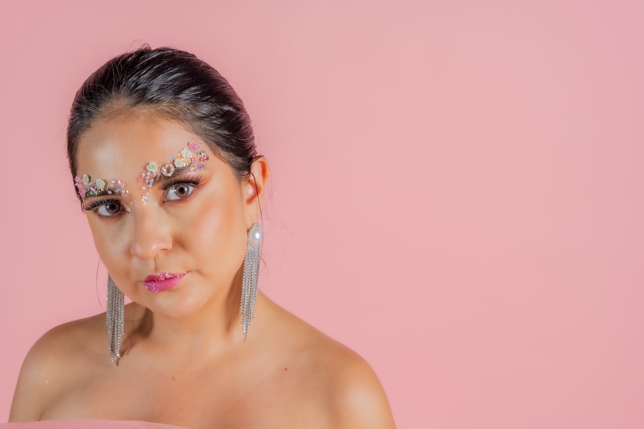 Stylish artistic portrait of a woman with creative makeup and elegant earrings against a pink background.