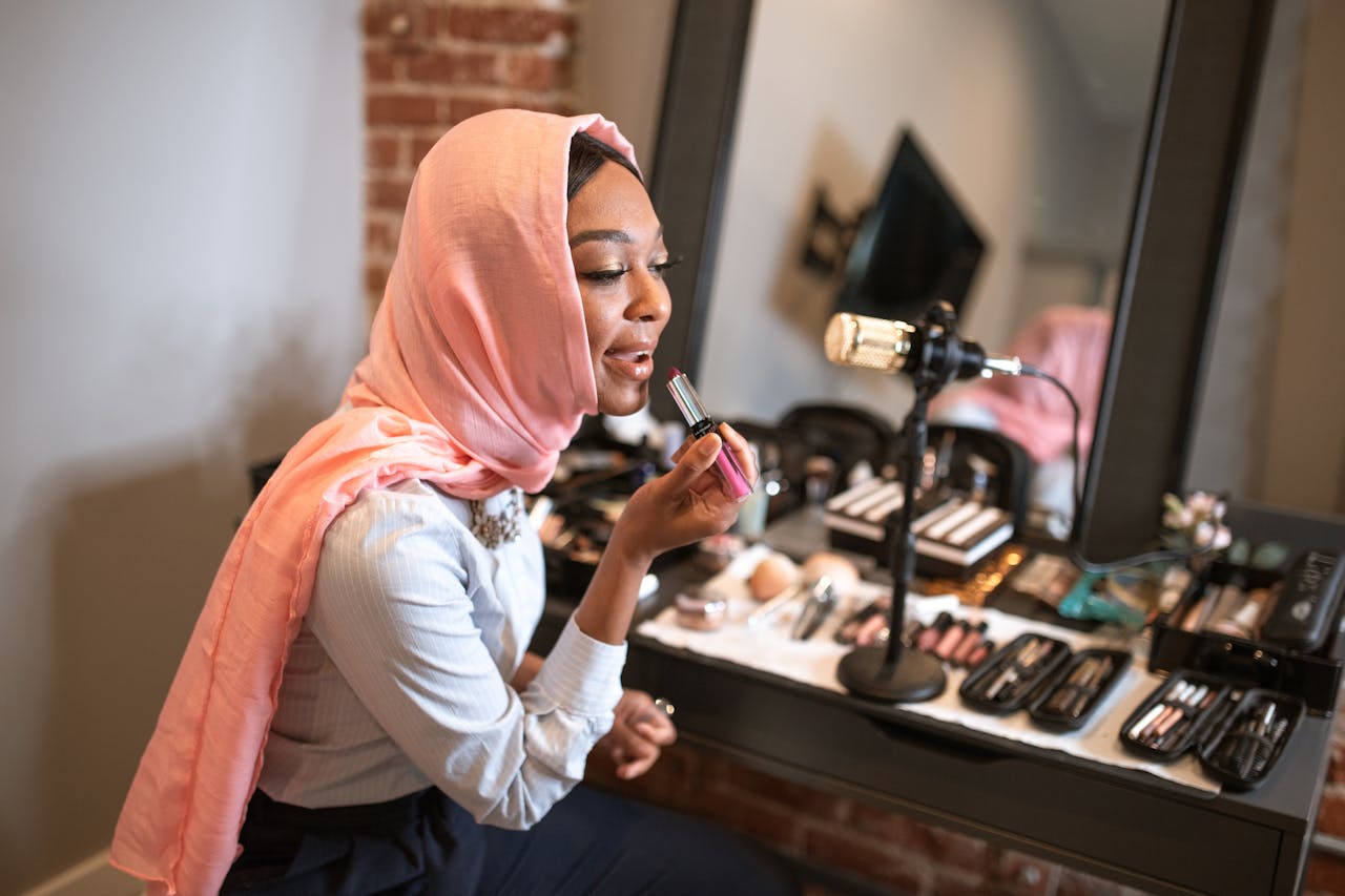 About Muslim woman in pink hijab applying makeup while recording a beauty vlog indoors.