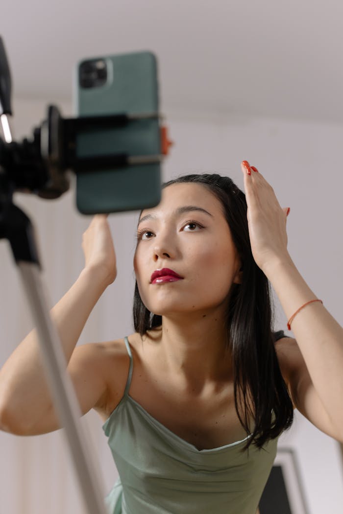 Services Asian woman adjusts hair and makeup in front of smartphone for live online content creation.