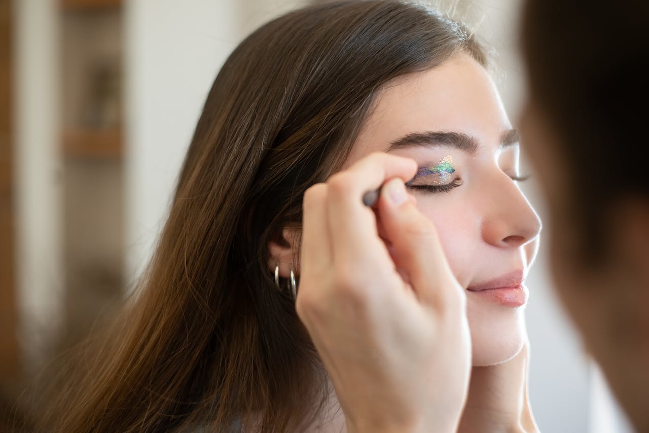 A close-up shot of a woman with eyes closed as a makeup artist applies glitter eyeshadow.