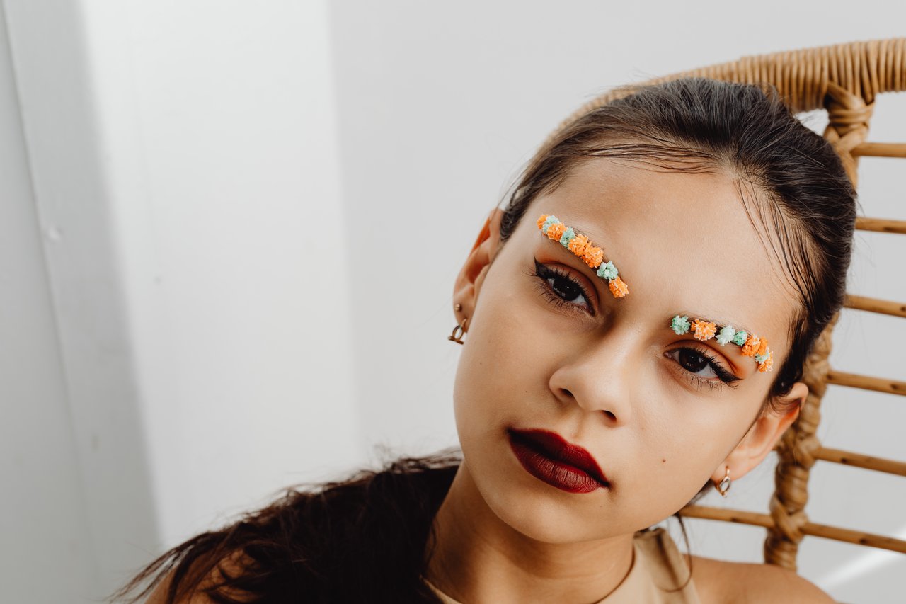 A young woman with artistic floral eyebrow makeup sitting indoors.